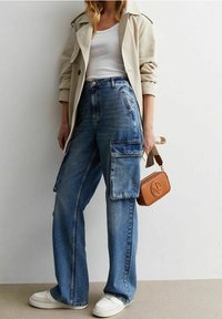 Trench coat de color claro sobre una camiseta blanca, combinado con jeans cargo de pierna ancha en un azul deslavado con grandes bolsillos laterales y zapatillas blancas.