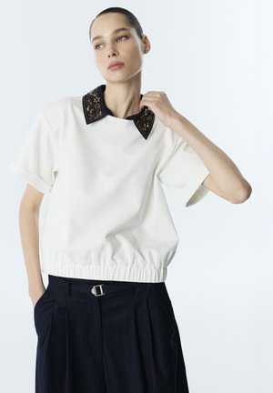 Vrouw draagt een wit shirt met korte mouwen met geborduurde zwarte kraag en zwarte broek, poseert met haar hand dichtbij de kraag tegen een effen achtergrond.