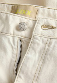 Μπεζ παντελόνι από τζιν με μερικώς ανοιχτό φερμουάρ, μεταλλικό κουμπί με χαραγμένο "JJXX" και υφασμάτινη ετικέτα με "JJXX" σε κίτρινο χρώμα.