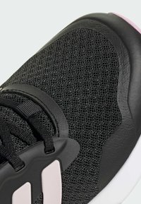 Scarpa sportiva nera con tomaia in rete testurizzata, dettagli neri lisci e un logo rosa chiaro. I lacci sono neri con un fermacorda in plastica.