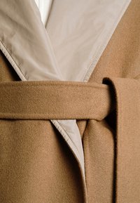 Manteau marron avec un col beige brillant et une ceinture. Le tissu est texturé et chaud, avec un design épuré et une légère brillance sur le col.