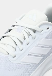 Gros plan d'une basket blanche Adidas avec des lacets et le logo signature à trois bandes sur une tige en mesh.