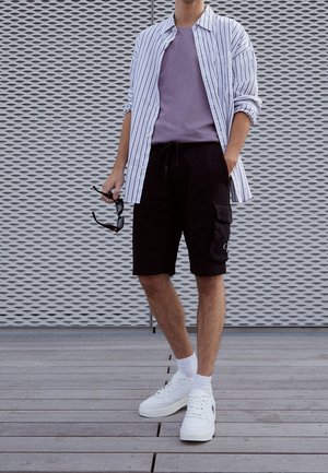 Pantaloni cargo neri con tasche laterali, t-shirt viola e camicia bianca a righe con bottoni. Sneakers bianche con suole spesse e calzini bianchi corti.