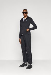 Giacca nera con zip e taglio aderente, abbinata a pantaloni neri e calzature coordinate. Gli accessori includono occhiali da sole scuri.