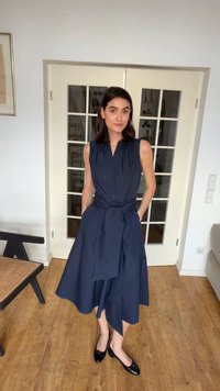 Frau steht drinnen, trägt ein ärmelloses marineblaues Midikleid mit gebundener Taille und schwarze Ballerinas, Hände in den Taschen, heller Holzboden.