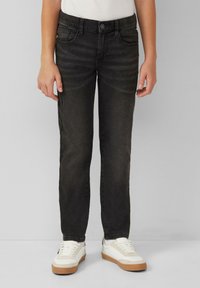 Bambino che indossa jeans slim-fit neri e sneaker bianche, in piedi contro uno sfondo grigio uniforme.