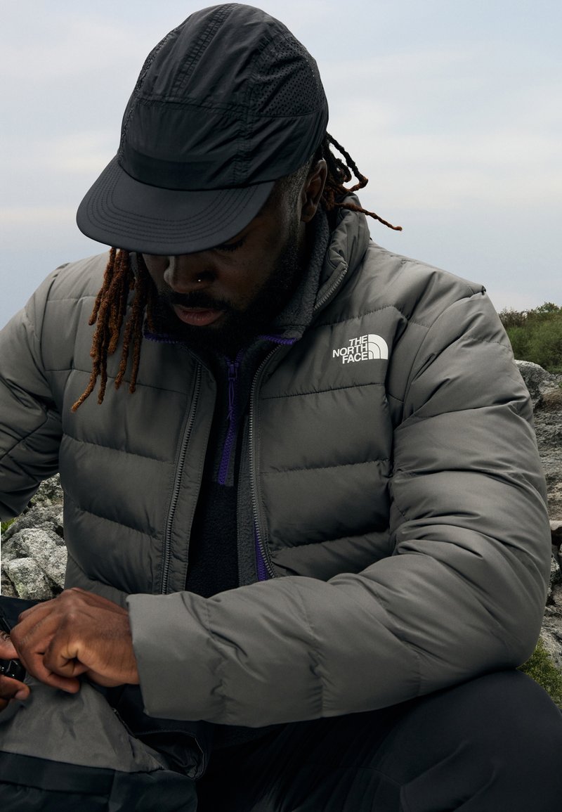 Grå pufferjakke med glidelås foran, lilla detaljer og "The North Face"-logo. Svart caps. Temaet sitter ved steiner og justerer en veske.