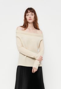 Suéter bege com os ombros à mostra, textura canelada, mangas longas e ajuste descontraído, combinado com uma saia plissada preta.