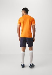 Oranž sportlik T-särk, lühikesed varrukad kontrastsete kantidega, koos mereväe siniste lühikesed pükstega. Valged põlvikud ja mustad tossud roheliste detailidega.