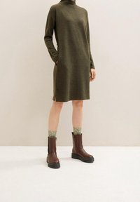 Olivi vihreä neulemekko korkealla kauluksella, sivutaskuilla ja halkioilla. Ruskeat nilkkurit mustilla pohjilla. Kiiltävät vihreät sukat näkyvissä.
