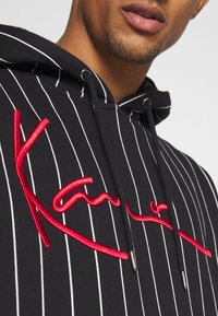 Mustaraidallinen huppari, jossa on punainen kirjailtu logo. Hupussa on metallipäällysteiset nyörit ja pehmeä pinta.