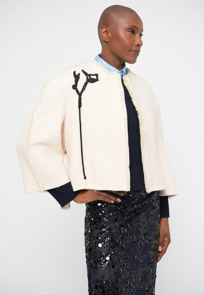 Balta, struktūrizuota trumpa striukė, pagaminta iš tekstūruotos medžiagos, su juodais gėlių siuvinėjimais priekyje, derinama su blizgančia sijonu.