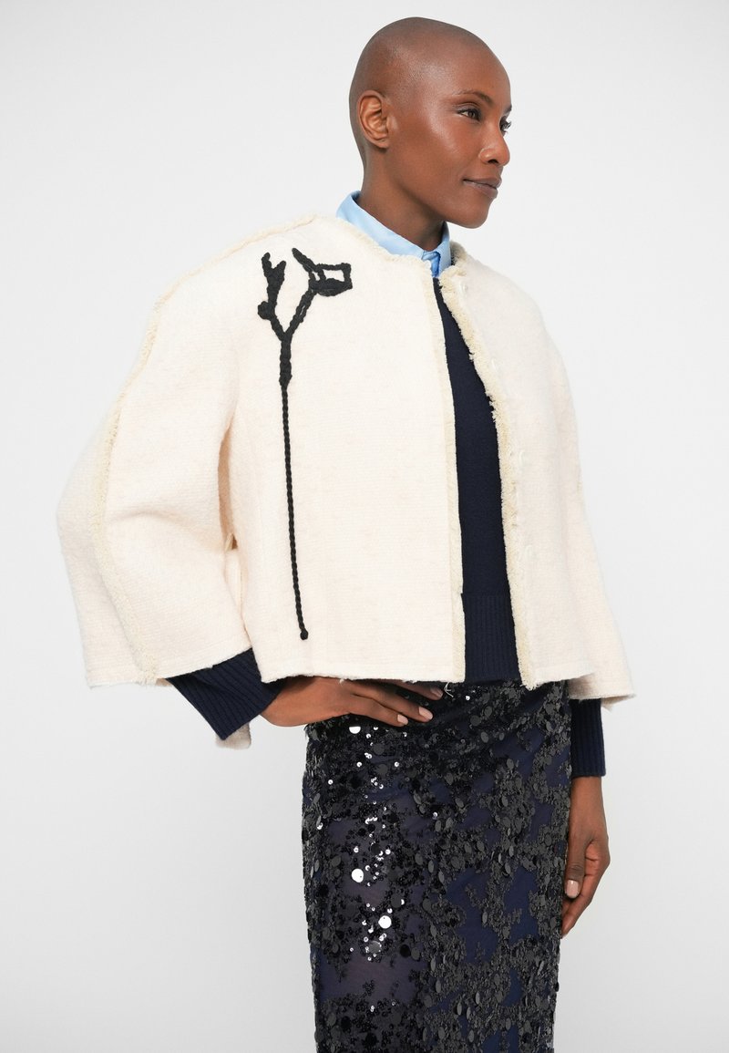 Giacca corta bianca, strutturata, realizzata in tessuto texture, con un dettaglio di ricamo floreale nero sul davanti, abbinata a una gonna di paillettes.