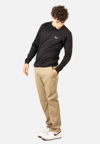 Schwarzes Langarmshirt mit weißem Logo, kombiniert mit beigen Hosen und weißen Sneakers mit roten Akzenten, in einer entspannten Pose stehend.