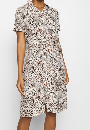 Femme portant une robe mi-longue à manches courtes avec un motif animalier en beige, noir et blanc, présentant une ceinture nouée à la taille et une petite fente à l'avant.