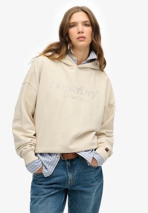Beige hoodie met een logo, gelaagd over een lichtblauw en wit gestreept shirt, gecombineerd met losse denim jeans met een riem.