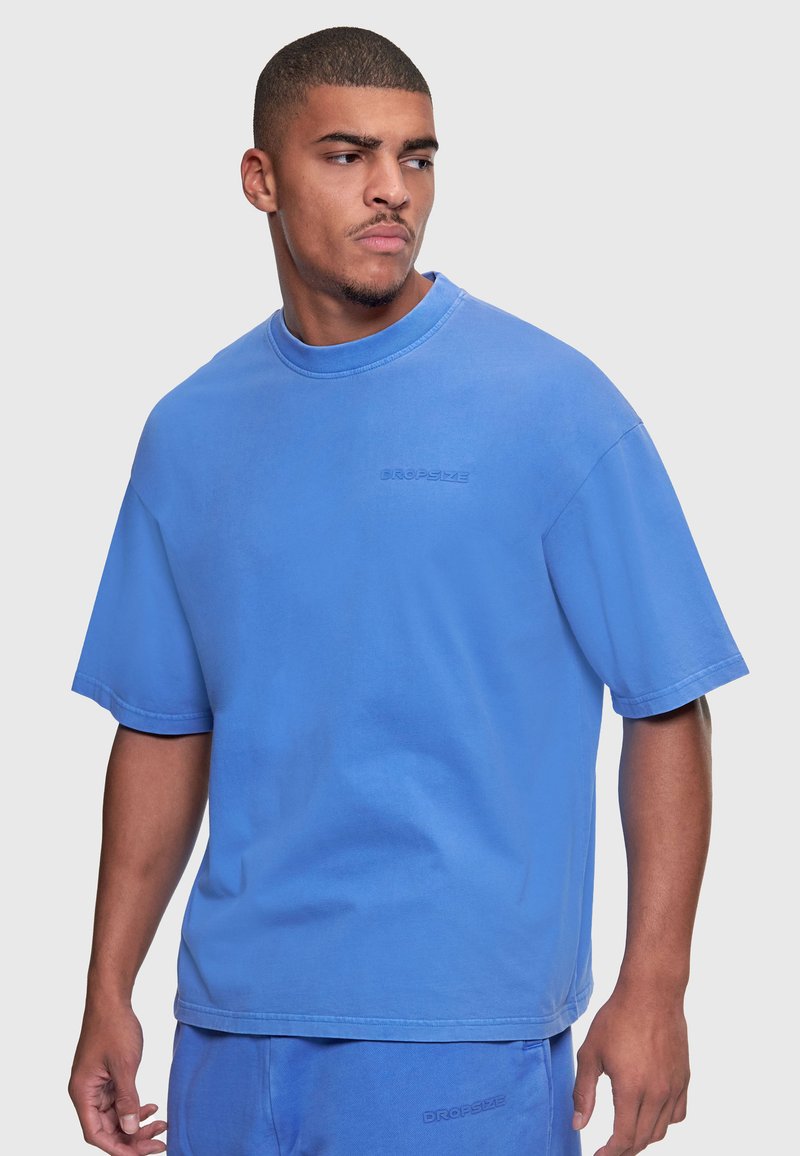 Homme portant un t-shirt bleu oversize assorti et un short, regardant vers sa gauche sur un fond clair uni.