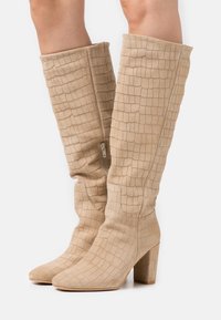 Bottes hauteur genou beiges avec un motif texturé de crocodile. Dotées d'un talon bloc épais et d'une fermeture éclair latérale pour un enfilage facile. Fabriquées en matière suède douce.