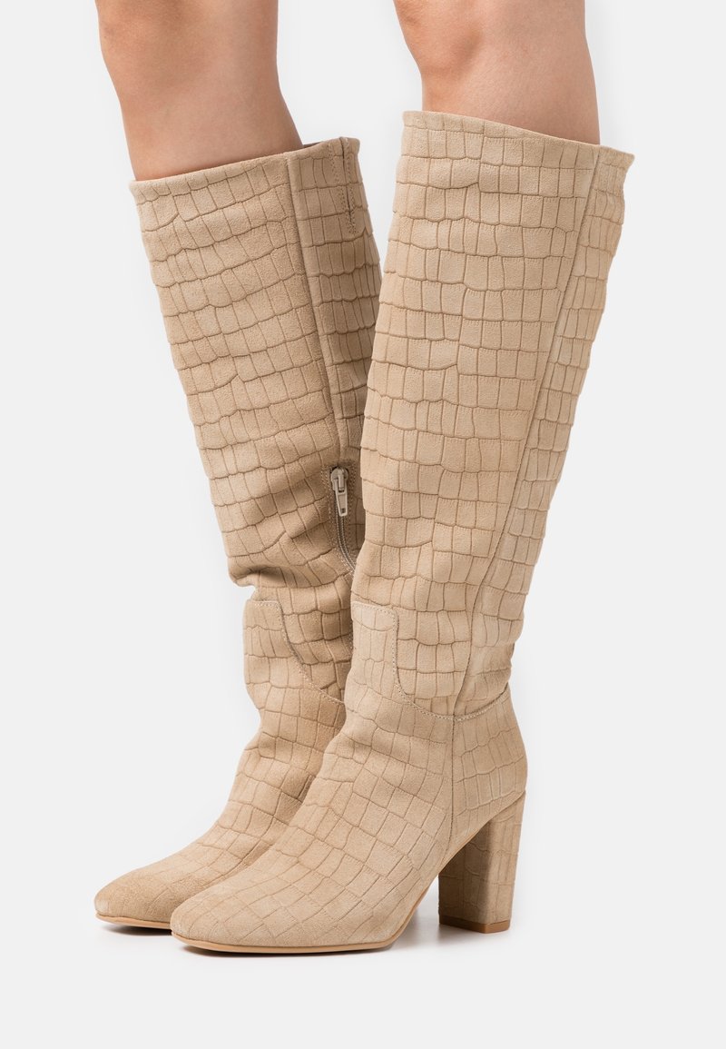 Bottes hauteur genou beiges avec un motif texturé de crocodile. Dotées d'un talon bloc épais et d'une fermeture éclair latérale pour un enfilage facile. Fabriquées en matière suède douce.