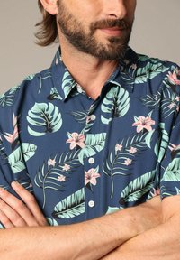 Korte mouw overhemd met knopen in marineblauw met groene tropische bladeren en roze bloemen. Heeft een kraag en een relaxte pasvorm.