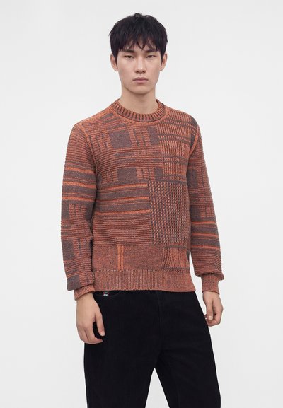 Jeune homme portant un pull en maille orange et marron à motifs avec un pantalon noir, debout devant un fond clair uni.