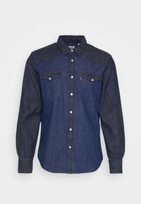 Camisa de mezclilla azul oscuro con mangas largas, frente abotonado, dos bolsillos en el pecho y un tiro de hombro en contraste. Botones de presión plateados.