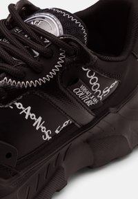 Baskets noires Versace Jeans Couture avec motif chaîne blanc, lacets noirs, couture en zigzag blanche et semelle épaisse texturée.