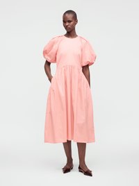 Kvinne iført en rosa midikjole med puffete korte ermer og flate spisse mørke sko, stående med hendene i lommer mot en ensfarget bakgrunn.