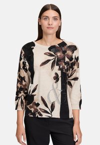 Femme portant un pull à motifs floraux beige et noir avec des manches trois-quarts et un pantalon noir, se tenant devant un fond uni.