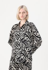 Svart och krämfärgad zebra-mönstrad skjorta med långa ärmar, krage och knappslutning framtill. Mjuk tyg med en flytande, avslappnad passform.