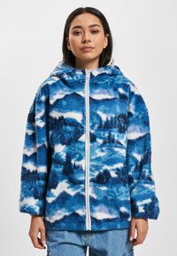 Blå fleece hettegenser med et fjellandskapsmønster i varierende nyanser av blått og hvitt. Full zip-lukking og hette.
