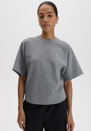 Vrouw die een loszittende grijze korte mouwen top en een zwarte broek draagt, staand tegen een eenvoudige lichte achtergrond.
