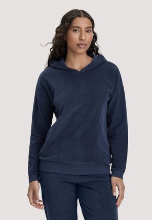 Frau mit langen lockigen Haaren, die einen dunkelblauen Kapuzenpullover und eine passende Hose trägt, vor einem schlichten hellen Hintergrund stehend.