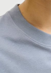 Κοντινό πλάνο του γιακά ενός γαλάζιου φούτερ με στρογγυλή λαιμόκοψη, που φοριέται από ένα άτομο με ανοιχτό καφέ δέρμα.