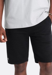Shorts noirs texturés avec un design côtelé, en coton, et un patch logo sur le côté, associés à une chemise gris clair.