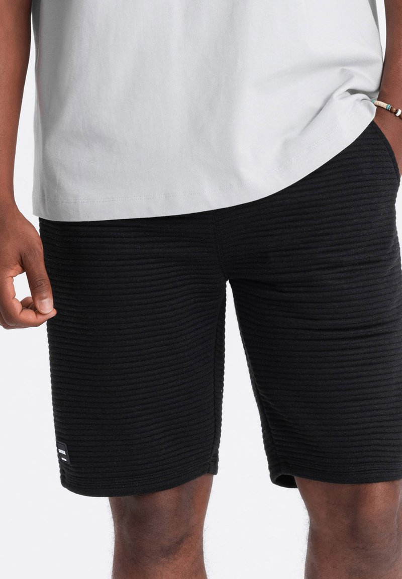 Shorts noirs texturés avec un design côtelé, en coton, et un patch logo sur le côté, associés à une chemise gris clair.