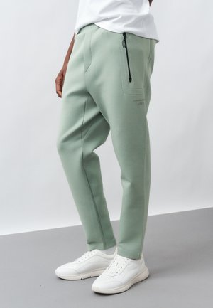 Άνδρας που φοράει ανοιχτό πράσινο παντελόνι με κωνική γραμμή και τσέπη με φερμουάρ, καθώς και λευκά αθλητικά παπούτσια, στέκεται σε γκρι δάπεδο με λευκή φόντο.