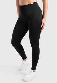 Leggings noirs taille haute en tissu doux et extensible avec une taille côtelée, associés à des chaussures de sport blanches.