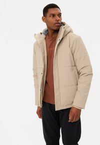 Giacca trapuntata beige con cappuccio, caratterizzata da un design a quadri e fodera grigia, indossata sopra una camicia marrone, abbinata a pantaloni scuri.