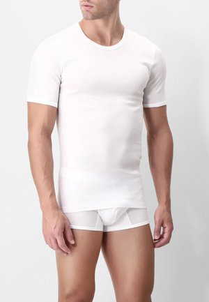 Uomo che indossa una maglietta bianca a maniche corte aderente e boxer coordinati bianchi su uno sfondo bianco uniforme.