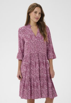 Kobieta o długich brązowych włosach, ubrana w różowo-białą sukienkę z wzorem, sięgającą do kolan, z rękawami 3/4 i dekoltem w serek, stojąca na jednolitym tle.