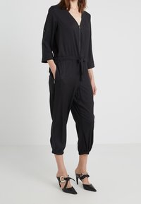 Jumpsuit noir avec une fermeture éclair à l'avant, col en V, manches 3/4, poignets élastiqués et ceinture à nouer. Assorti à des escarpins à bout pointu avec un détail de boucle.