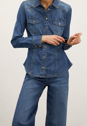 Femme portant une chemise en denim bleu avec des poches avant et un jean en denim assorti taille haute à jambes larges, ajustant le poignet de la chemise.