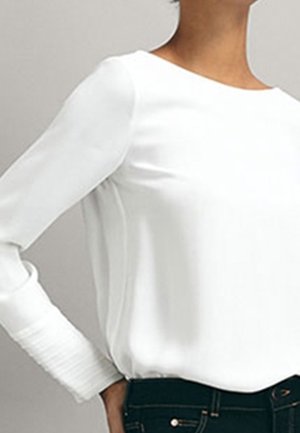 Vrouw die een wit blouse met lange mouwen en geplooide manchetten draagt, gecombineerd met een donkere spijkerbroek, afgebeeld van nek tot taille tegen een effen achtergrond.