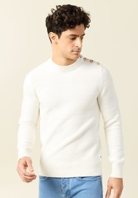Weißer Pullover aus Strick mit strukturierten Streifen, geripptem Ausschnitt und Bündchen. Verfügt über dekorative Knöpfe an der linken Schulter, kombiniert mit blauen Jeans.