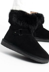 Botas negras de ante hasta el tobillo con un suave cuello de piel sintética, que cuentan con un detalle de correa lateral y una suela de goma texturizada.