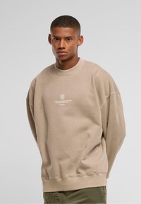 Beige oversized sweatshirt med rund hals, ribbede manchetter og kant. Har teksttryk foran i hvid og blødt, tekstureret stof.