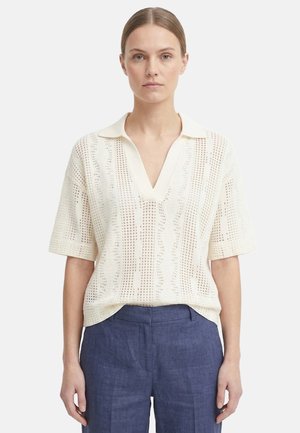 Donna che indossa una polo a maniche corte in maglia color crema con scollo a V e pantaloni blu a vita alta, in piedi su uno sfondo semplice.