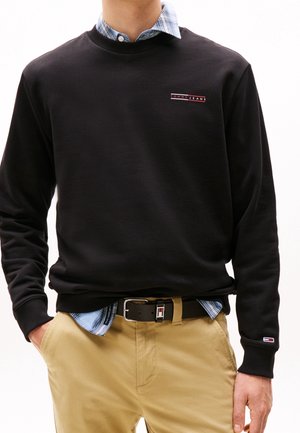Osoba ubrana w czarną bluzę Tommy Jeans na niebieską koszulę w kratę oraz beżowe spodnie z czarnym paskiem, ręce w kieszeniach.