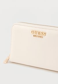 Világos bézs bőr pénztárca aranyszínű "GUESS" logóval, cipzáras záródással és látható varrással. Cipzárhúzó füllel rendelkezik márkajelzéssel.